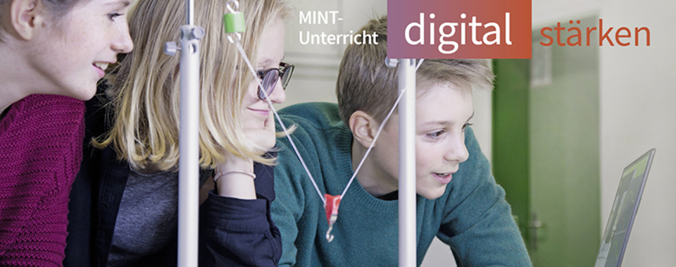 Das Bild zeigt drei Schüler, die konzentriert an einem Experiment oder einer digitalen Präsentation arbeiten. Im Hintergrund sind Apparaturen sichtbar, die in einem MINT-Unterricht eingesetzt werden, um das Interesse an Naturwissenschaften zu fördern.