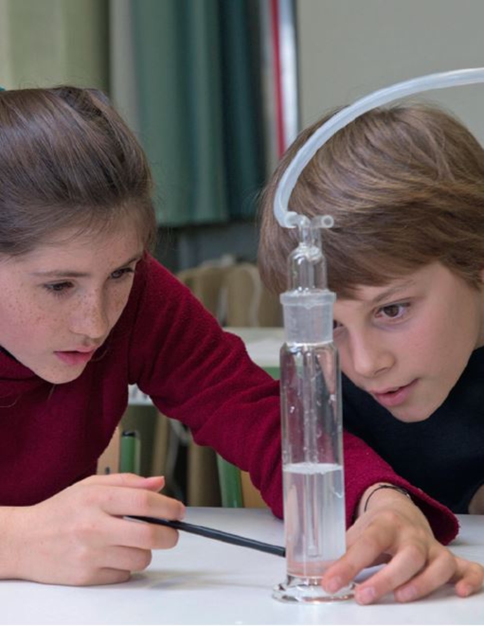 Das Bild zeigt zwei Kinder, die konzentriert an einem Experiment mit einemZylindermessgerät arbeiten. Sie beobachten den Wasserstand und verwenden ein spezielles Gerät, um den Vorgang zu steuern, was auf ein naturwissenschaftliches Experiment hinweist.