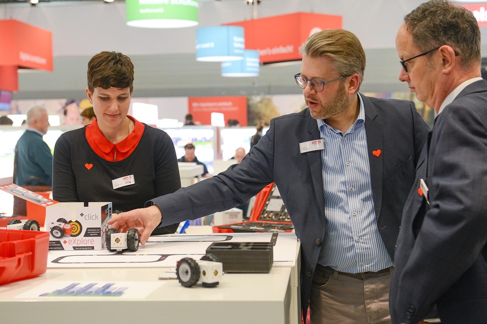 Das Bild zeigt einen Messestand von Cornelsen Experimenta. Zu sehen sind drei Personen, die sich über den Lernroboter eXperiBot austauschen.