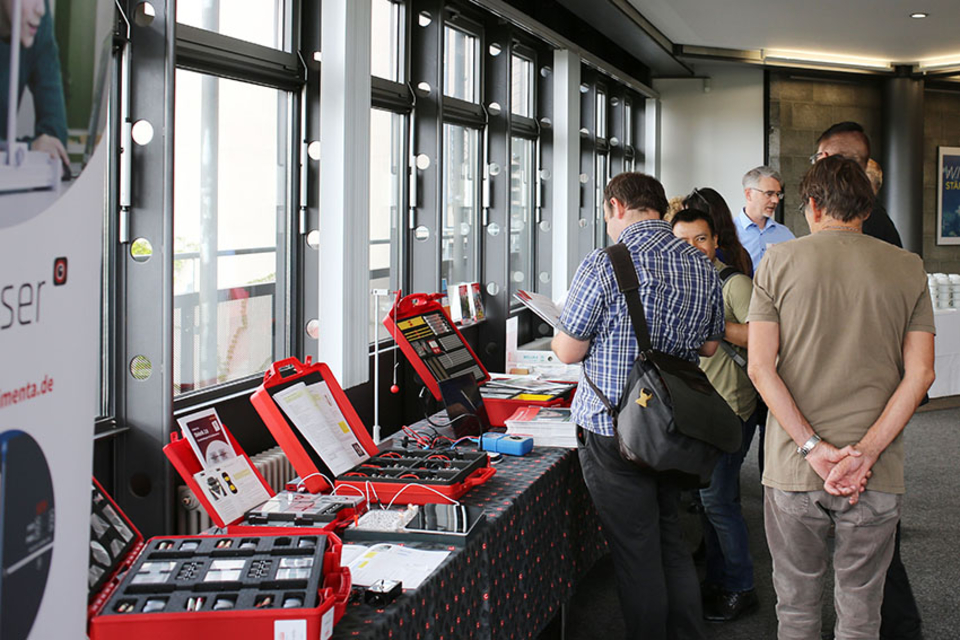 Das Bild zeigt eine Gruppe von Personen, die an einem Stand für Experimentierbedarf stehen, wo verschiedene Koffer und Materialien präsentiert werden. Der Stand ist hell erleuchtet und die Besucher scheinen sich für die angebotenen Produkte zu interessier