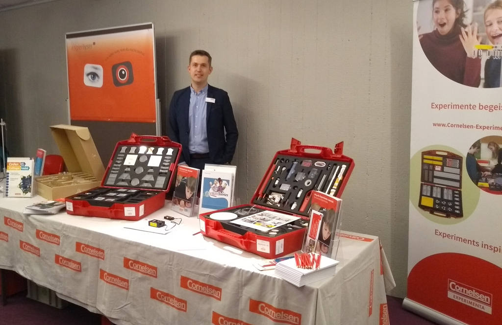 Das Bild zeigt einen Stand mit Experimentiermaterialien für das Bildungswesen, einschließlich zwei roter Koffer mit verschiedenen Experimentierwerkzeugen. Im Hintergrund steht ein Mann, der den Stand präsentiert.