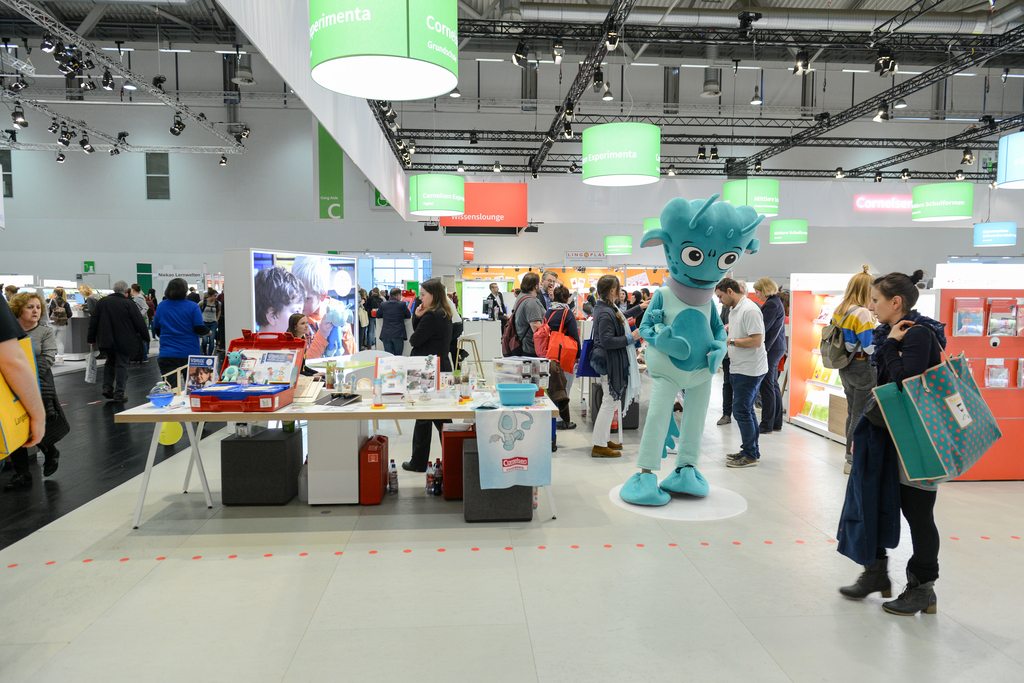 Das Bild zeigt einen Messestand mit einem großen blauen Maskottchen und mehreren Personen, die sich um Stände mit Experimentiermaterialien versammeln. Die Umgebung ist hell und modern, mit verschiedenen Displays und Informationsständen.