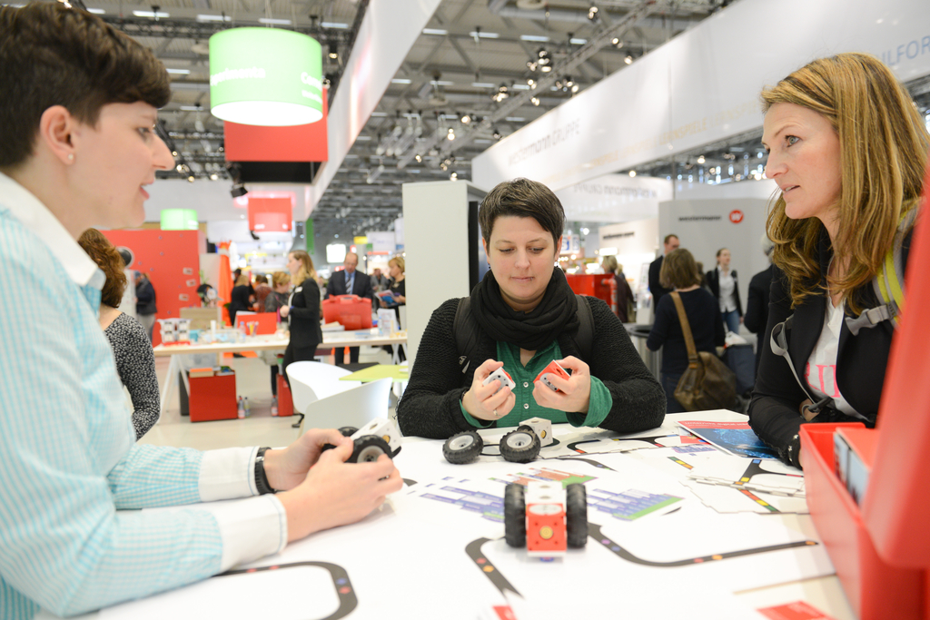 Das Bild zeigt drei Frauen, die an einem Tisch mit Experimentiermaterialien und arbeiten, darunter ist auch der Lernroboter eXperiBot. Im Hintergrund sind Stände und Besucher einer Messe zu sehen, was auf eine bildungsorientierte Veranstaltung hinweist.