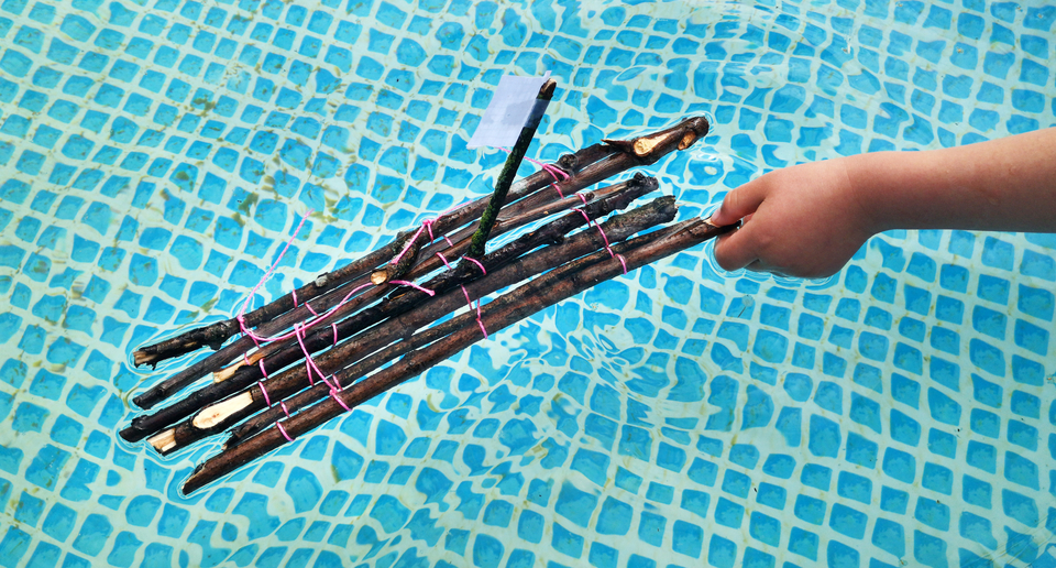 Das Bild zeigt ein Miniatur-Floß, das aus mehreren zusammengebundenen Ästen besteht und auf der Wasseroberfläche schwimmt. Eine Hand hält das Boot, während es in einem blauen Wasserbecken bleibt.