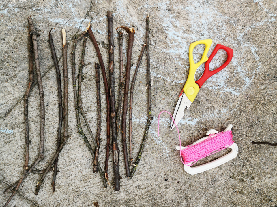 Das Bild zeigt eine Anordnung von mehreren dünnen Ästen auf einem grauen Untergrund, neben denen eine bunte Schere sowie ein Spulenhalter mit pinkem Faden liegen. Diese Materialien sind ideal für verschiedene kreative Experimente in der Bildung.