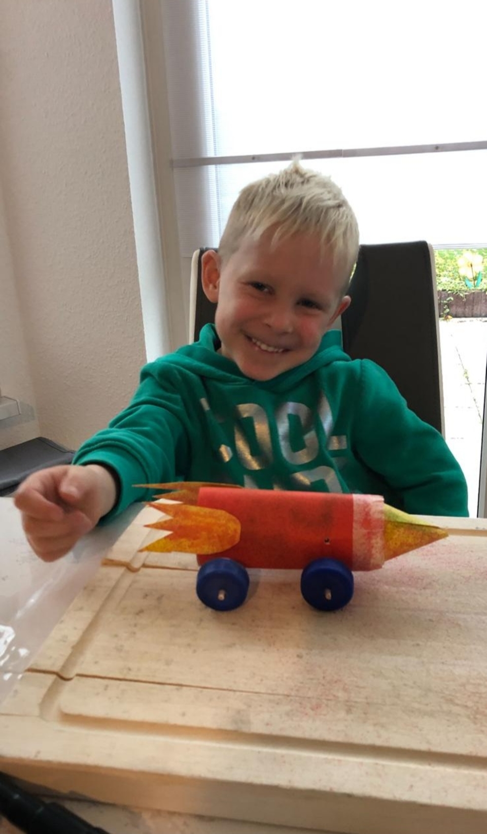 Auf dem Bild ist ein lächelnder Junge zu sehen, der stolz sein selbstgebautes Modell einer Rakete präsentiert. Die Rakete ist farbenfroh gestaltet und steht auf einer Holzplatte mit Rädern, während der Junge mit einer Hand darauf zeigt.