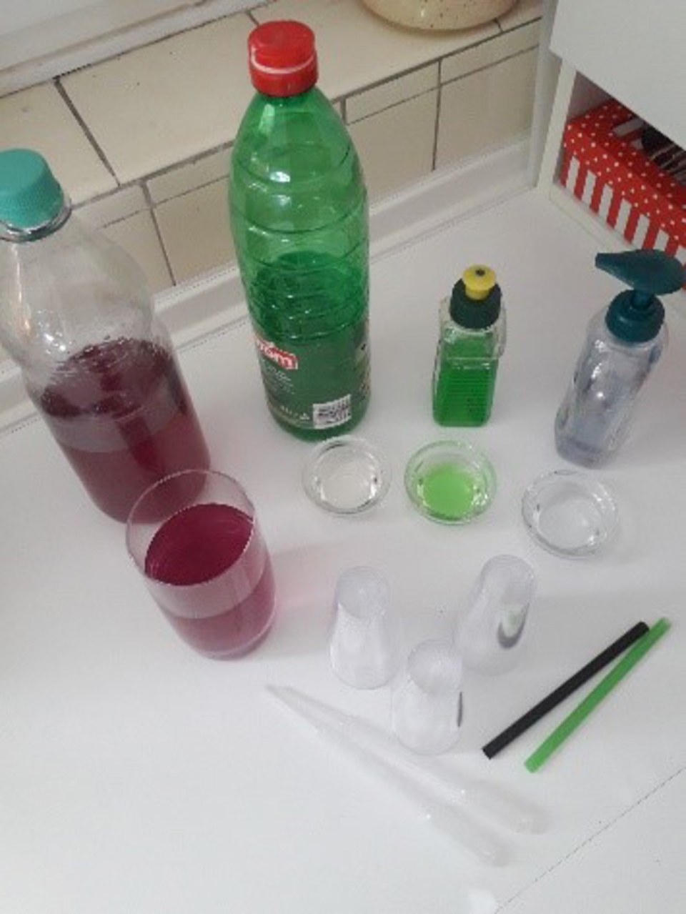 Das Bild zeigt eine Anordnung verschiedener Flüssigkeiten in Flaschen und Gläsern sowie einige Mess- und Experimentiergeräte, wie Pipetten und Bechergläser. Die Farben der Flüssigkeiten reichen von Rot über Grün bis zu Transparent, passend für chemische E