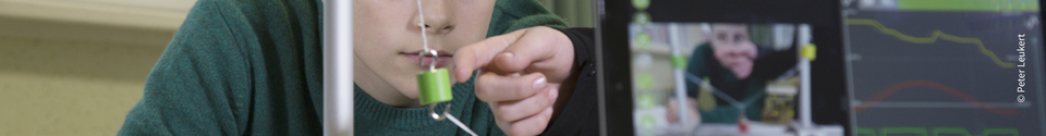 Das Bild zeigt einen Schüler, der konzentriert an einem Experiment arbeitet, bei dem er einen grünen Magneten manipuliert. Im Hintergrund ist ein Bildschirm zu sehen, der vermutlich Daten oder Ergebnisse des Experiments anzeigt.