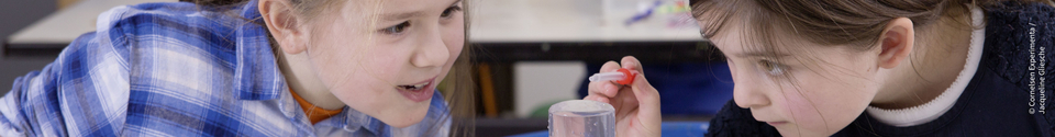 Auf dem Bild sind zwei Kinder zu sehen, die fasziniert auf ein Experiment in einem Becher schauen. Eines der Kinder hält eine Pipette und scheint bereit zu sein, etwas in die Flüssigkeit zu geben.