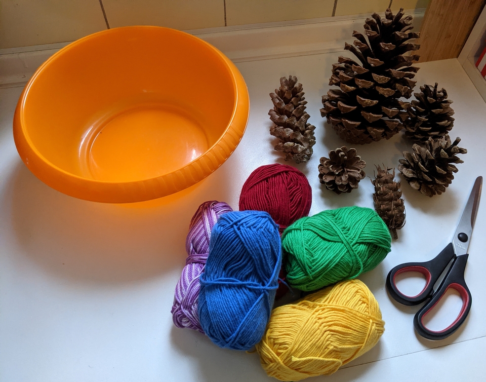 Ein orangefarbener Behälter steht neben verschiedenen Kiefernzapfen und bunten Wollknäueln in Blau, Grün, Rot, Lila und Gelb. Eine Schere liegt ebenfalls auf dem Tisch, was auf potenzielle Bastelaktivitäten hinweist.