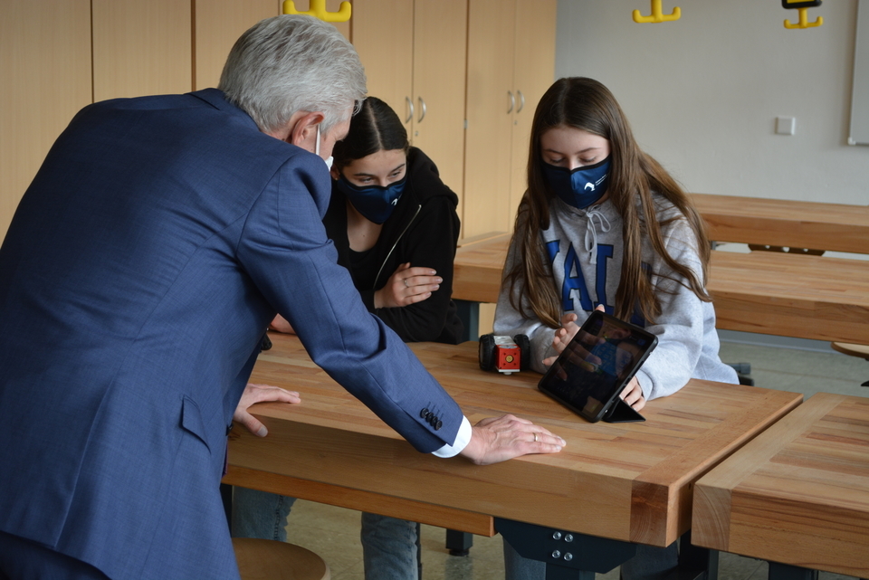 Das Bild zeigt einen Lehrer, der mit zwei Schülerinnen an einem Tisch arbeitet, während sie ein Tablet nutzen. Auf dem Tisch befindet sich ein kleiner Roboter, und alle drei Personen tragen Masken.