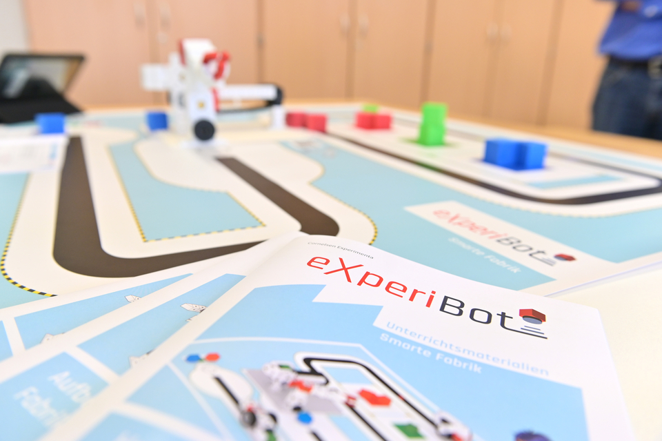 Das Bild zeigt einen Tisch mit einem Experimentier-Bereich, auf dem eine Strecke mit farbigen Blöcken und einem kleinen Robotermodell angeordnet ist. Vorne liegen Unterrichtsmaterialien mit der Aufschrift "eXperiBot - Smarte Fabrik".
