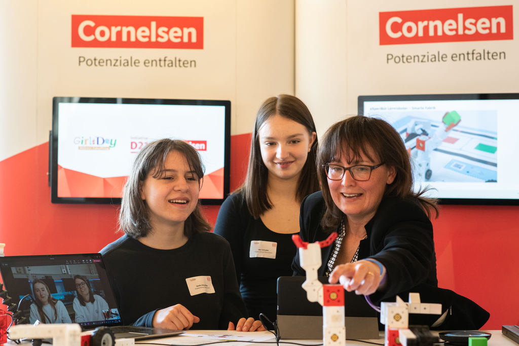 Das Bild zeigt drei Personen an einem Stand des Unternehmens Cornelsen, die sich mit dem eXperiBot Lernroboter beschäftigen. Im Hintergrund sind ein Laptop und Bildschirmanzeigen zu sehen, die auf Bildungsinitiativen hinweisen.
