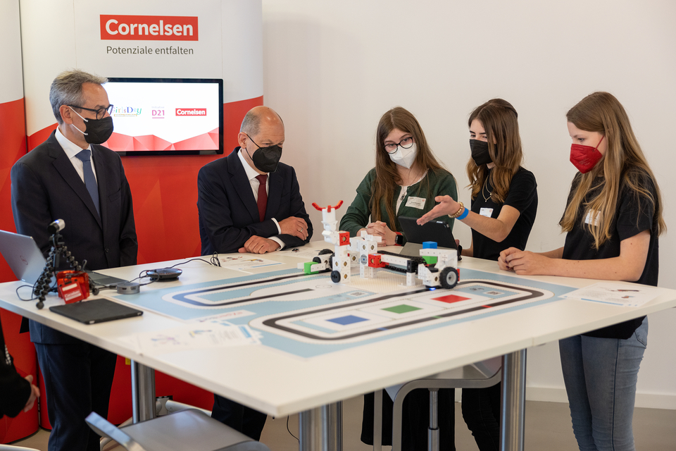 Das Bild zeigt eine Gruppe von Schülerinnen und dem Bundeskanzler, die an einem Tisch mit dem eXperiBot Lernroboter in der Smarten Fabrik diskutieren. Hinter ihnen steht ein Banner von Cornelsen, und sie diskutieren lebhaft über die technischen Möglichkei