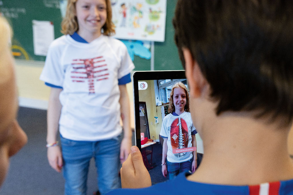 Das Bild zeigt ein Kind, das ein T-Shirt mit anatomischen Motiven trägt und vor einer Tafel steht. Ein anderes Kind hält ein Tablet, auf dem eine Augmented-Reality-Anwendung zu sehen ist, die die Anatomie des Trägers darstellt.