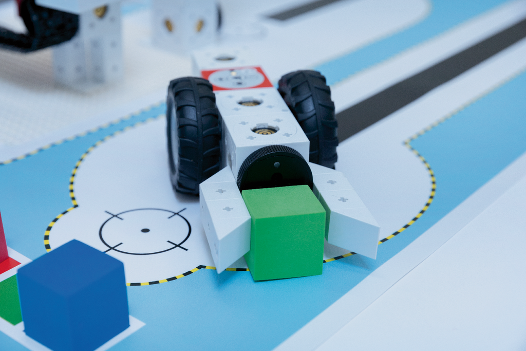 Das Bild zeigt den Lernroboter eXperiBot, der auf einer abgebildeten Lernumgebung platziert ist. Er führt eine Aufgabe aus, bei der er einen grünen Block aufnimmt und sich dabei zwischen bunten Formen bewegt.