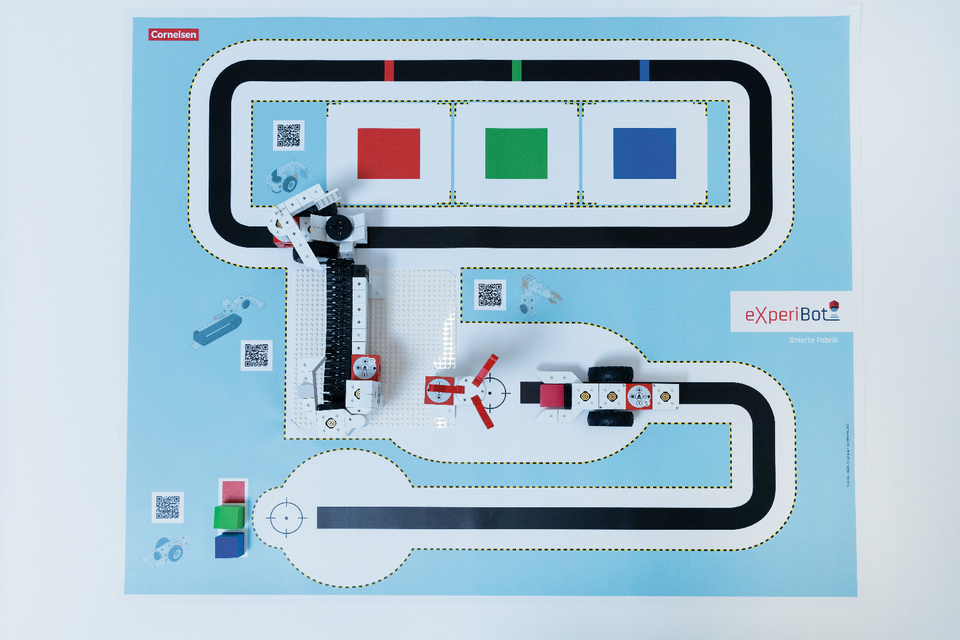 Das Bild zeigt eine farbige Spielmatte mit einer komplexen Straßenführung und verschiedenen farbigen Feldern, die für Experimente mit Robotern genutzt werden. In der Mitte ist ein Roboterarm sowie eine kleine Fahrzeugstruktur zu sehen, die für Bildungsprojekte konzipiert sind.