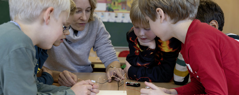 Das Bild zeigt eine Lehrerin, die mit einer Gruppe von Kindern im Sachunterricht an einem Experiment arbeitet. Sie konzentrieren sich auf eine interessante Aktivität, bei der elektrische Komponenten und Drähte auf einem Tisch angeordnet sind.