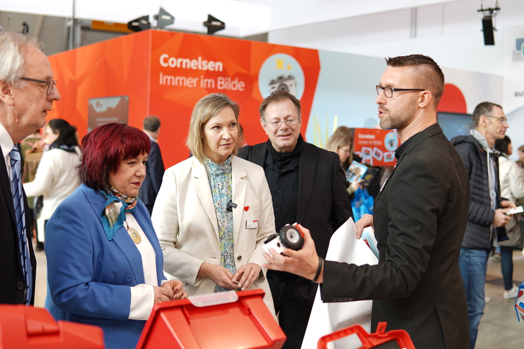 Das Bild zeigt eine Gruppe von Personen, die einem Präsentierenden aufmerksam zuhören, während er ihnen ein Produkt zeigt. Im Hintergrund ist ein auffälliges, orangefarbenes Banner von Cornelsen zu sehen, das auf deren Engagement im Bildungsbereich hinweist.