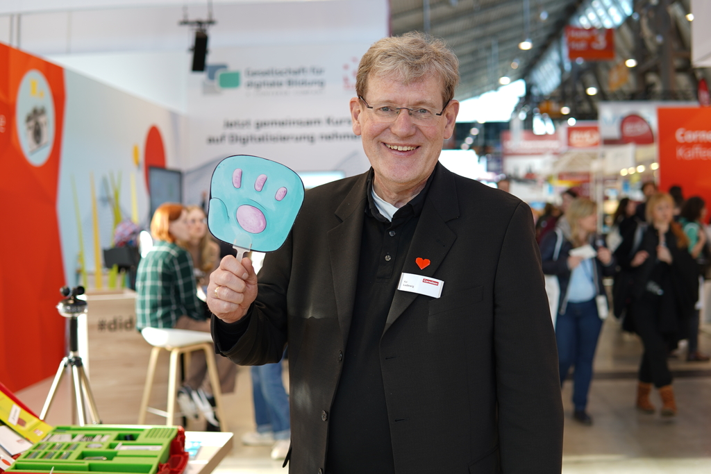 Das Bild zeigt einen lächelnden Mann in einem schwarzen Blazer, der ein plakatiertes Hand-Symbol hält. Im Hintergrund sind Menschen und Stände einer Bildungsmesse zu sehen, was auf eine Veranstaltung für experimentelle Lernmaterialien hindeutet.