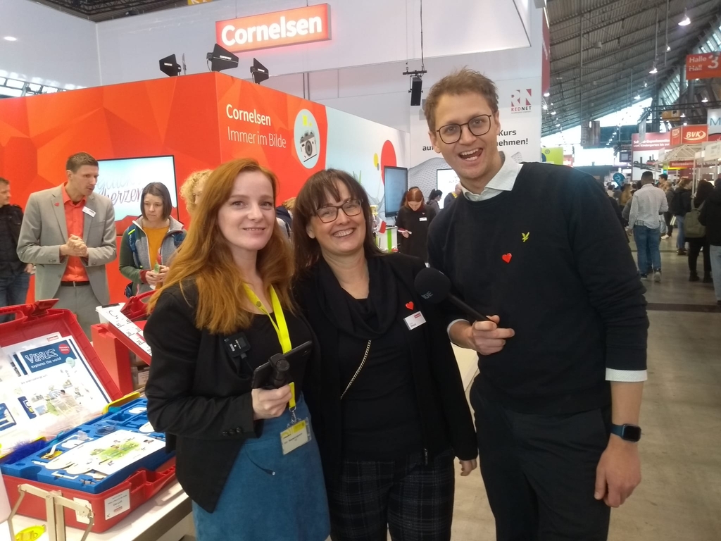 Das Bild zeigt mehrere Personen an einem Messestand von Cornelsen, der sich mit Experimentierbedarf für das Bildungswesen beschäftigt. Im Vordergrund lächeln zwei Frauen und ein Mann in formeller Kleidung in die Kamera, während im Hintergrund weitere Besu