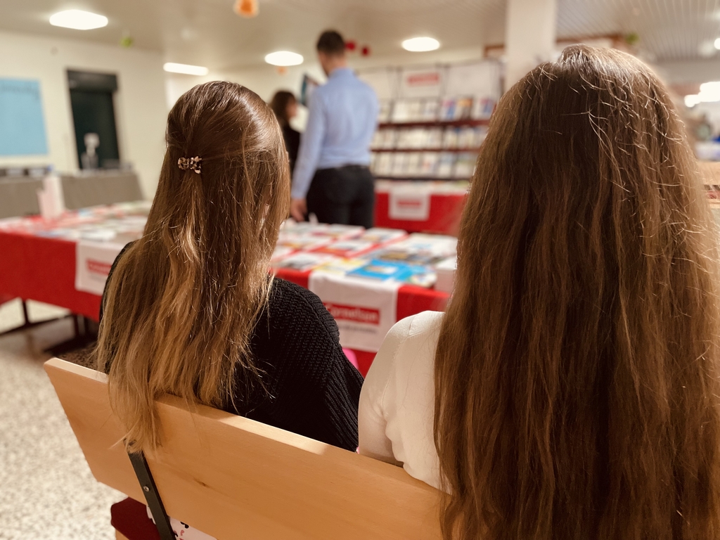 Das Bild zeigt zwei Schülerinnen, die auf einer Bank sitzen und sich den Cornelsen-Stand mit Bücher- und Experimentiermaterialien im Hintergrund anschauen. Dieser Stand ist im Rahmen einer Veranstaltung für das deutsche Bildungswesen aufgebaut.