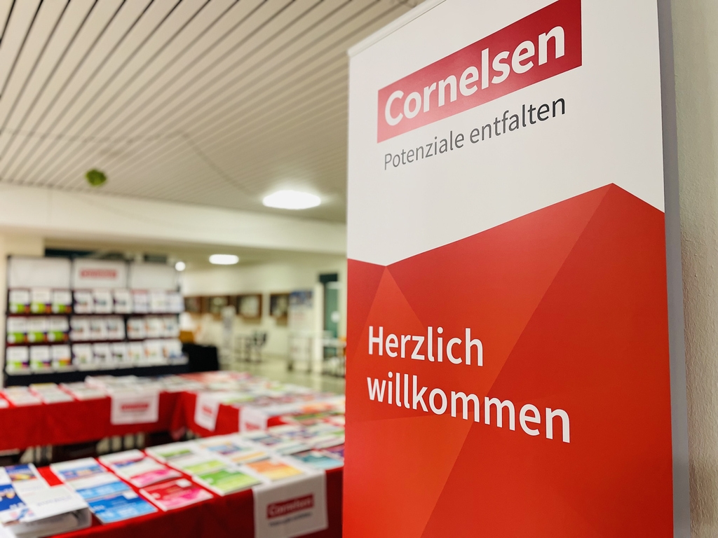 Das Bild zeigt einen Aufsteller von Cornelsen mit der Aufschrift "Herzlich Willkommen". Im Hintergrund sind Regale mit Büchern und anderen Materialien für den Unterricht zu sehen.