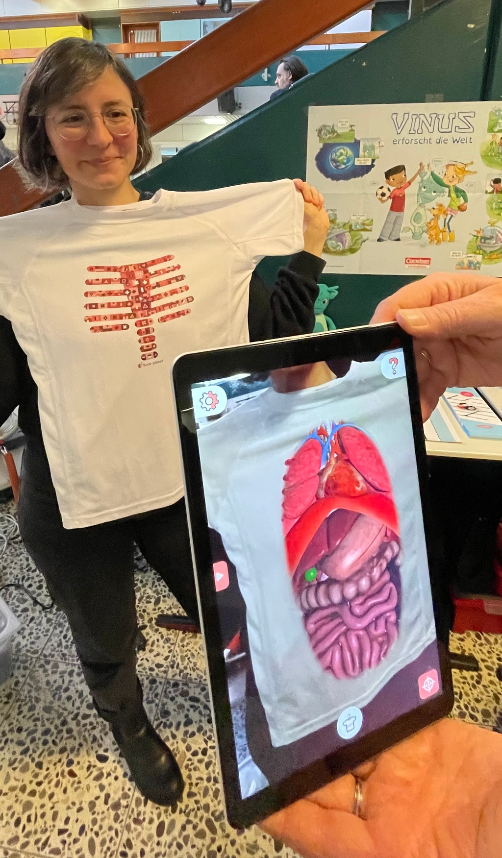 Auf dem Bild hält eine Frau das Magic T-Shirt mit einem anatomischen Print, der menschliche Rippen zeigt, vor ihren Oberkörper. Daneben wird auf einem Tablet mithilfe der Magic T-Shirt-App eine erweiterte Realität (AR)-Darstellung des Inneren eines menschlichen Körpers gezeigt.