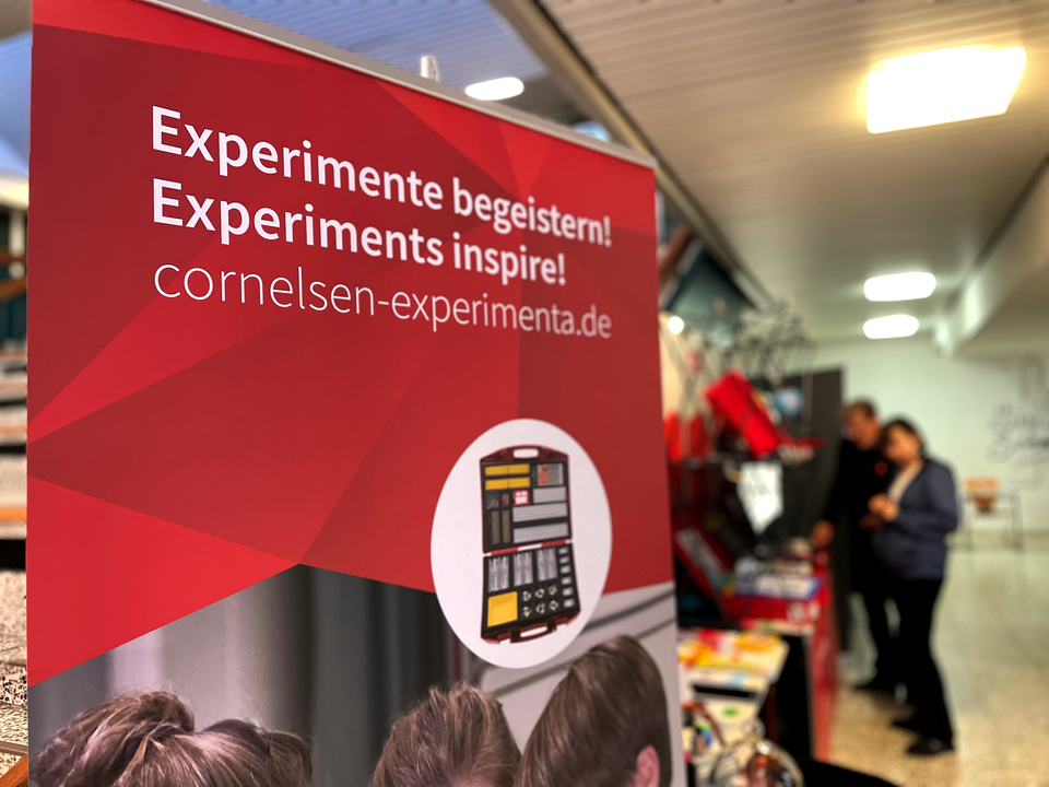 Das Bild zeigt einen Banner von Cornelsen Experimenta auf der MNU Bremerhaven. Es handelt sich um einen Stand für Experimentierbedarf mit der  Botschaft „Experimente begeistern!“ Im Hintergrund stehen zwei Personen und betrachten das Sortiment.