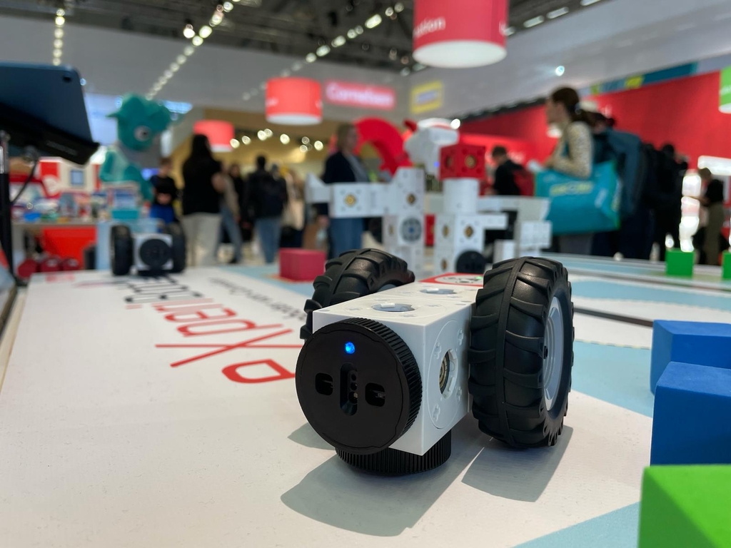 Das Bild zeigt eine Nahaufnahme des eXperiBot Explorers, einem kleinen, weißen Roboter mit großen Gummireifen, der auf einer Farbfläche in einem modernen Setting positioniert ist. Im Hintergrund sind Besucher der didacta 2024 und weitere Stände zu sehen.