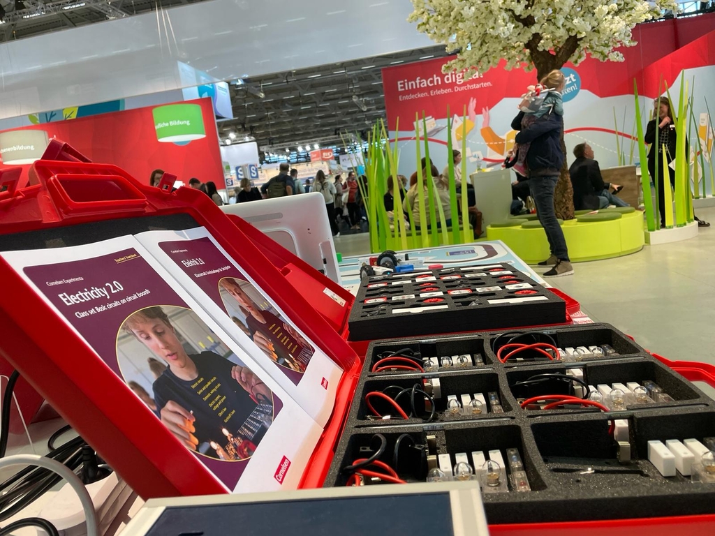 Das Bild zeigt eine rote Kunststoffbox mit Lehrmaterialien zum Thema Elektrizität, darunter zwei Bücher und verschiedene elektronische Bauteile wie Kabel und Lämpchen. Im Hintergrund sind Besucher einer Bildungsveranstaltung zu sehen.
