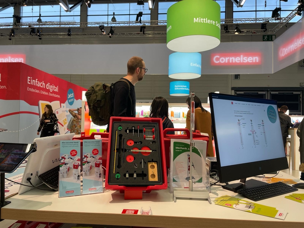 Das Bild zeigt einen Messestand von Cornelsen Experimenta mit Produkten für die Sekundarstufe, darunter der Rote Koffer "Mechanik 2.0" im Vordergrund sowie einem Computer, der ein Experiment zeigt. Im Hintergrund sind farbige Leuchtschilder.