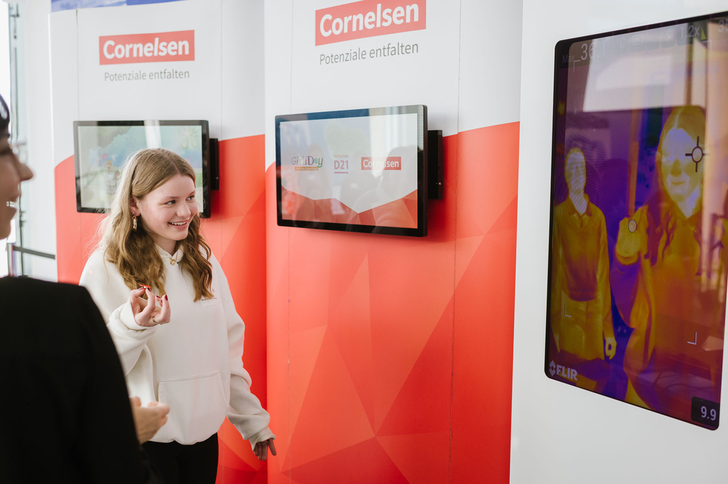 Girls_Day_2024_Schuelerin_Wärmebildkamera / Ein Mädchen in einem weißen Hoodie lächelt und zeigt auf ein Display mit einer Wärmebildaufnahme. Das Display zeigt das Cornelsen-Logo und den Slogan "Potenziale entfalten". Im Hintergrund befinden sich weitere 