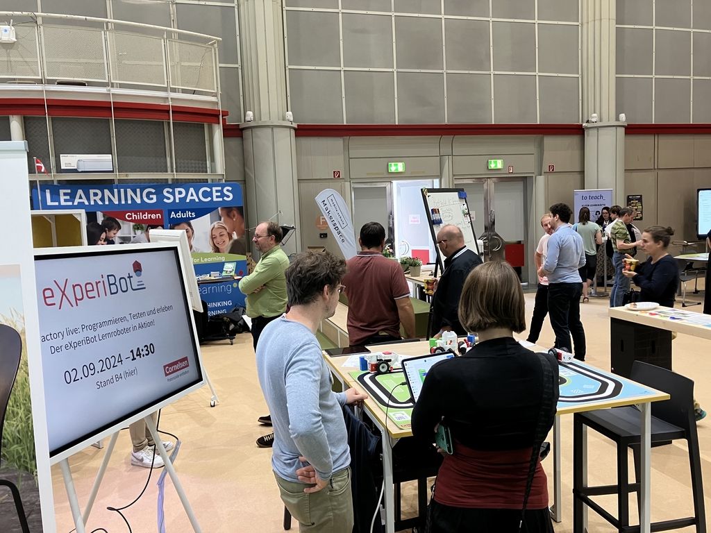 Mobile Schule Tagung 2024, im Vordergrund ist ein Messestand mit aufgebautem Lernroboter eXperiBot, im Hintergrund sind Menschen.