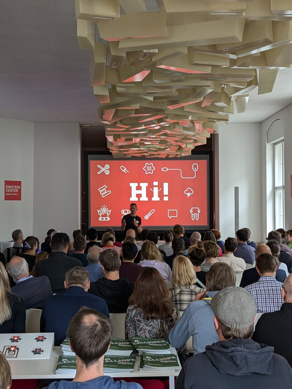 Das Bild zeigt eine Veranstaltung in einem modernen Raum, wo ein Sprecher vor einem großen Bildschirm mit der Begrüßung "Hi!" präsentiert. Im Vordergrund sind Teilnehmer zu sehen, während auf Tischen verschiedene Materialien und Informationsblätter liegen