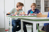 Das Bild zeigt zwei Jugendliche in einem Klassenzimmer, die an einem Experimentiergerät arbeiten. Der junge Mann links konzentriert sich auf einen Versuch, während der andere mit seinem Handy beschäftigt ist.
