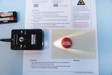 Das Bild zeigt ein Experimentiergerät mit Laser- und LED-Funktion, das auf einem Arbeitsblatt zur Durchführung von Lichtversuchen positioniert ist. Daneben liegt ein runder Marker mit dem Logo „Cornelsen Experimenta“, der das Licht des Lasers reflektiert.
