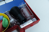Das Bild zeigt ein Set von Experimentierbedarf, das aus verschiedenen Glasbehältern, einem schwarzen Messzylinder und einem Röhrchen mit einer chemischen Substanz besteht. Zudem sind farbige Gummischnüre in einer gelben Schale enthalten, die für Experimen