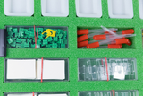 Das Bild zeigt eine bunte Anordnung von Experimentiermaterialien auf grünem Schaumstoff. Es sind grüne Bauteile, rote Pipetten, transparente Behälter und eine kleine gelbe Komponente sichtbar, die alle für Bildungszwecke verwendet werden können.