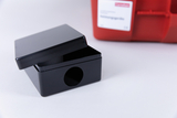 Das Bild zeigt eine schwarze, quadratische Box mit Deckel und einer Öffnung an der Seite. Im Hintergrund ist ein rotes Behältnis mit einem Etikett zu sehen, das auf Experimentierbedarf hinweist.