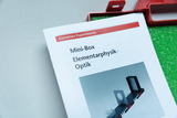 Das Bild zeigt eine Anleitung für eine Mini-Box zur Elementarphysik mit dem Schwerpunkt Optik, herausgegeben von Cornelsen Experimenta. Auf der Seite sind verschiedene optische Elemente abgebildet, die für Experimente genutzt werden können.