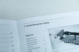 Das Bild zeigt eine Seite eines Handbuchs mit dem Titel "Ausbreitung des Lichtes". Es listet Materialien für ein Experiment auf und zeigt eine Abbildung eines Handexperiments mit einer Taschenlampe und einem lichtstreuenden Objekt.