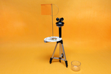 Das Bild zeigt einen experimentellen Apparaturaufbau mit einem Kompass auf einem Stativ, einem Pendel und einer orangefarbenen Fahne. Daneben befindet sich ein transparenter Behälter, der vermutlich für die Durchführung von Experimenten verwendet wird.