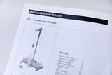 Das Bild zeigt eine Abbildung aus einem Lehrbuch zur Mechanik fester Körper, die einen Versuchsaufbau zur Bestimmung des Volumens eines Körpers darstellt. Neben der Grafik sind die benötigten Materialien für das Experiment aufgelistet.
