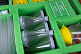 Das Bild zeigt eine sortierte Werkzeugbox mit verschiedenen Experimentiermaterialien, darunter transparente Reagenzgläser, Mikroskopträger und wasserfeste Folien. Die Teile sind in einem grünen Schaumstoffpolster sicher angeordnet, um Ordnung und Schutz z