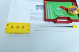 Das Bild zeigt ein Experimentier-Set zur Untersuchung von Schall, bestehend aus einem gelben Element mit Löchern, verschiedenen Röhren und einem roten Behälter mit Zubehör. Dazu liegt eine Anleitung, die die Durchführung der Experimente erläutert.