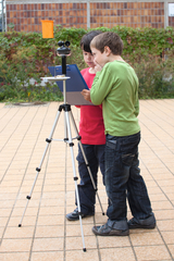 Das Bild zeigt zwei Kinder, die an einem stabilen Stativ mit einem Messgerät und einem orangefarbenen Fähnchen arbeiten. Sie schauen auf ein Clipboard, um ihre Ergebnisse festzuhalten und scheinen sich intensiv über ihr Experiment auszutauschen.