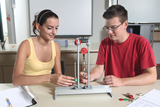Das Bild zeigt zwei Schüler, die an einem Experimentiergerät arbeiten, das für Physik- oder Technikunterricht im Bildungswesen verwendet wird. Der Junge und das Mädchen interagieren mit verschiedenen Gewichten und einem Aufhängesystem, während sie aufmerk
