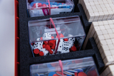 Das Bild zeigt eine Vielzahl von Lernspielmaterialien, darunter Spielsteine, Würfel und farbige Plättchen, die ordentlich in durchsichtigen Behältern aufbewahrt sind. Diese Materialien sind in einem schaumstoffgepolsterten Container arrangiert, der für de