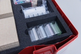 Das Bild zeigt eine rote Aufbewahrungsbox, die mit Schaumstoffeinsätzen gefüllt ist. In den Fächern sind verschiedene transparenten Behälter mit unterschiedlichen Materialien sowie einige Kartonstücke angeordnet, die wahrscheinlich für Experimentierzwecke
